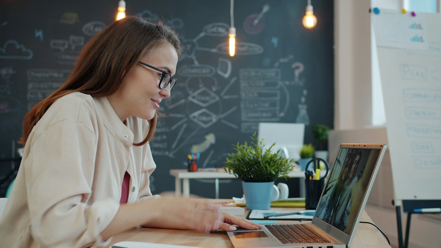 Woman video conferencing on a laptop at a desk with a chalkboard wall.